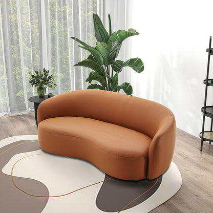 The Porter - Comfortable Sofa, with its modern curved brown leather design, sits on a patterned rug in a bright room with wood floors, beside a large green plant and a side table with flowers by sheer-curtained windows.
