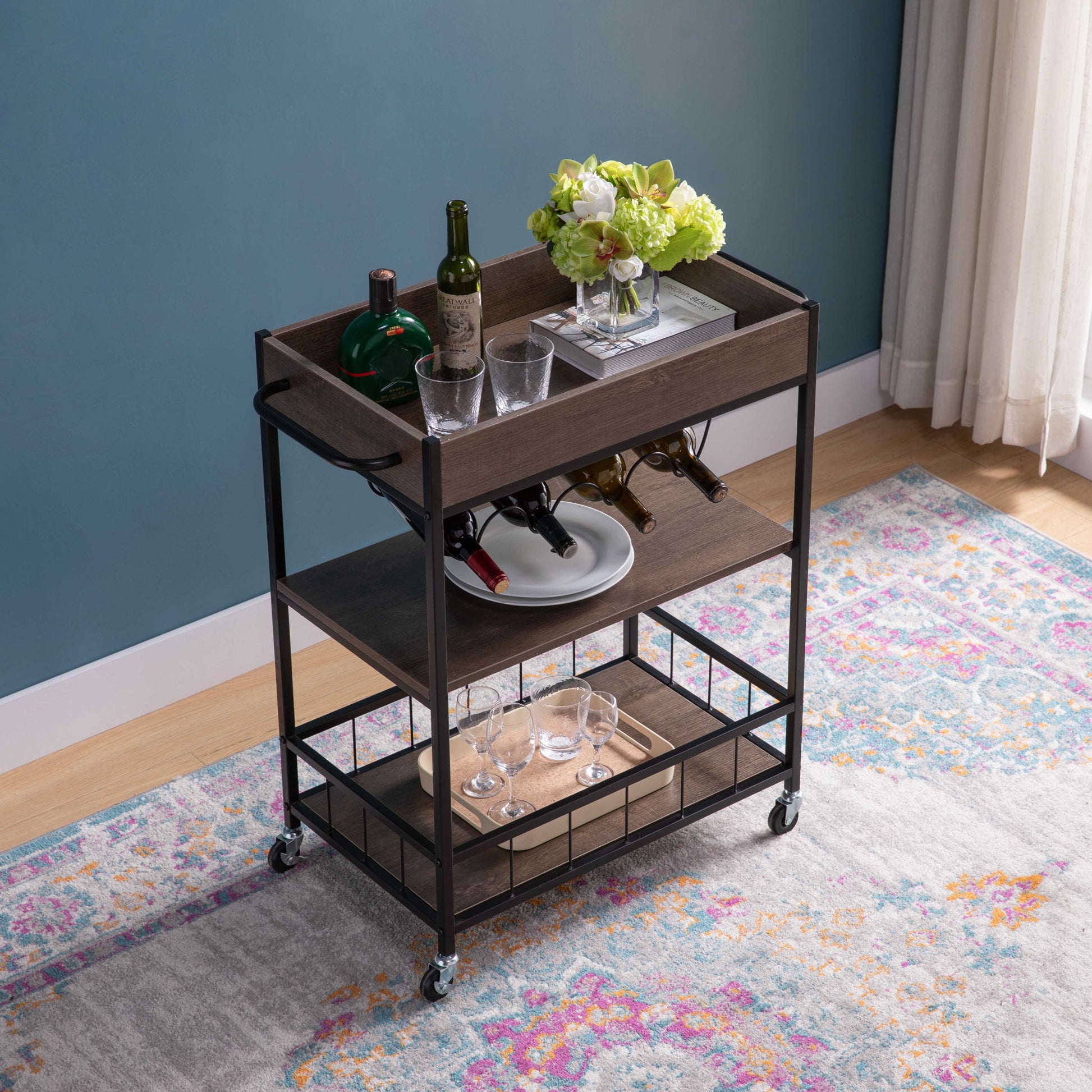 The Rolling Kitchen Cart With Storage And Four Wine Bottle Rack, featuring metal frames, wooden shelves, and small wheels, is set on a colorful rug in a blue-walled room with light curtains. Bottles, glasses, and flowers decorate the cart.