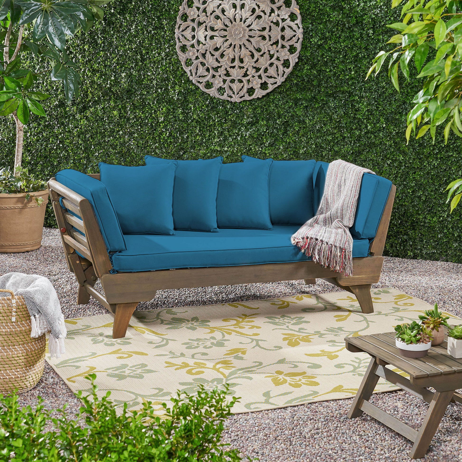 The Ottavio Convertible Sofa Daybed with water-resistant cushions and a gray striped throw blanket sits on a floral rug, surrounded by green plants and lattice wall décor on a garden patio.