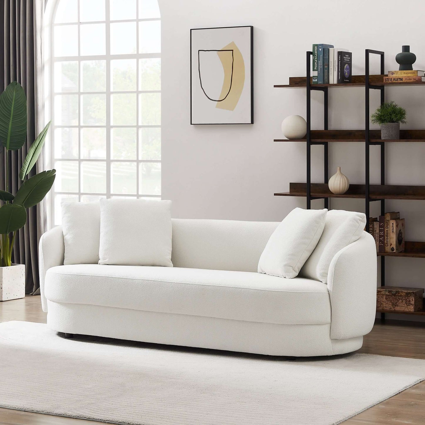 A modern living room with the Dylan - Modern French Boucle Sofa, three white cushions, a black open shelving unit with decor, a large window with curtains, a potted plant, abstract wall art, and a white rug on wood flooring.