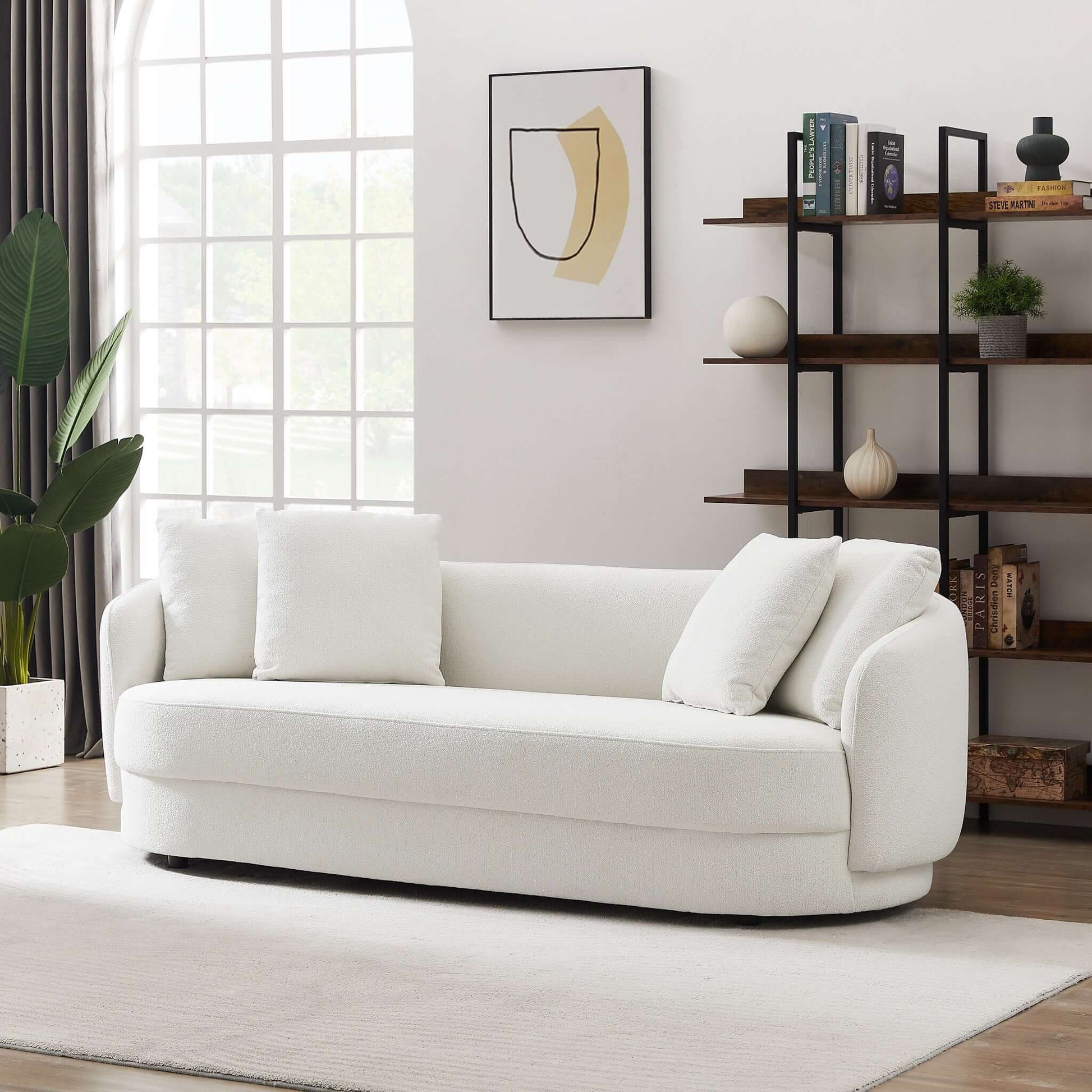 A modern living room with the Dylan - Modern French Boucle Sofa, three white cushions, a black open shelving unit with decor, a large window with curtains, a potted plant, abstract wall art, and a white rug on wood flooring.