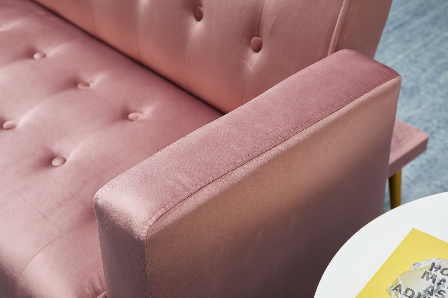 Close-up of the Comfort Compact Design Sofa Bed in pink velvet with button tufting, focusing on the armrest and seat. A white round table with a yellow object and gray item appears in the corner.