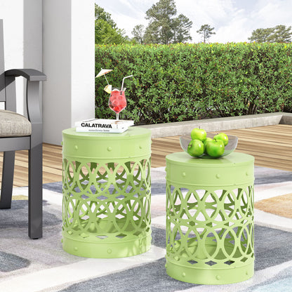Two Outdoor Metal S/2 End Tables with circular cut-out patterns are on a patio rug—one displaying green apples in a glass bowl, the other holding a book, sunglasses, and a red cocktail. Lush bushes and trees form the backdrop.