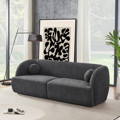 A modern living room featuring the Anna - French Boucle Sofa in dark gray, two round pillows, a black floor lamp, abstract black and white art, large windows with white curtains, and a green potted plant.