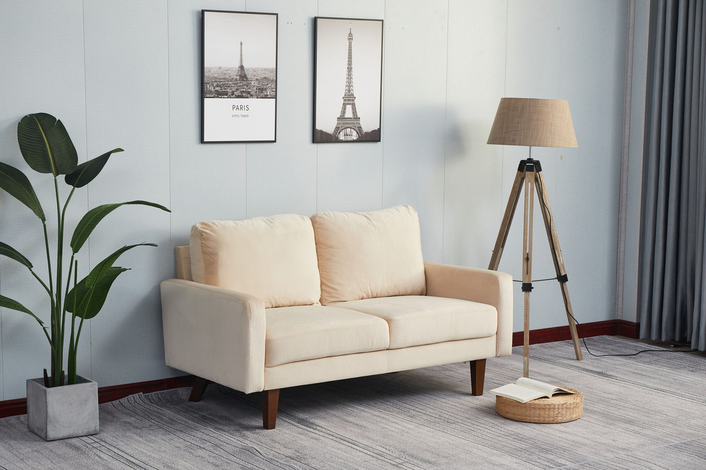 A Comfortable Sofa With Durable Construction sits on a gray rug in a modern living room, alongside a tall potted plant, two framed Eiffel Tower prints, a tripod floor lamp, and an open book on the floor.