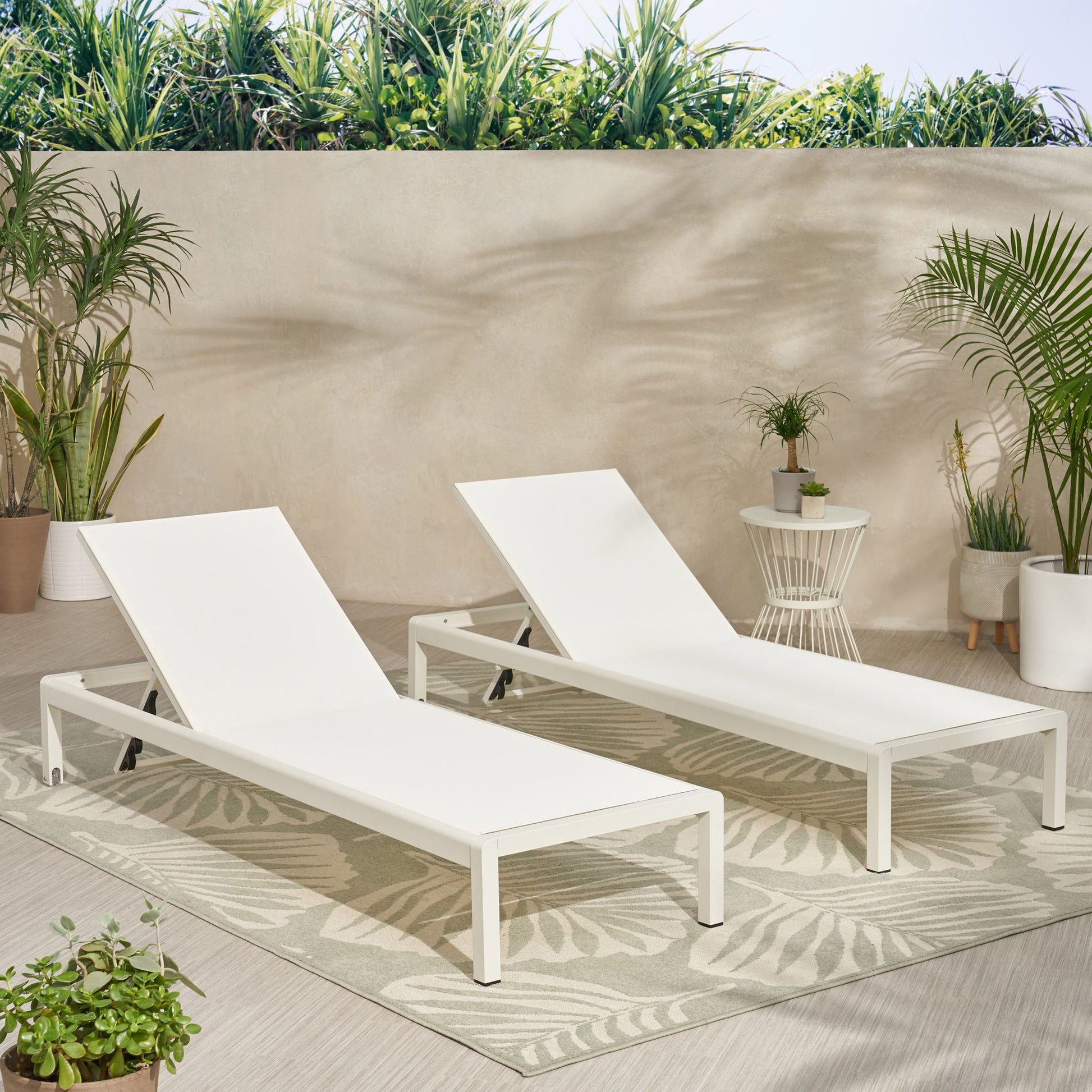 Two Outdoor Patio Chaise Lounges with 5 Reclining Positions sit side by side on a patterned outdoor rug, surrounded by potted plants against a light stone wall with green foliage above. The setup feels bright and inviting.