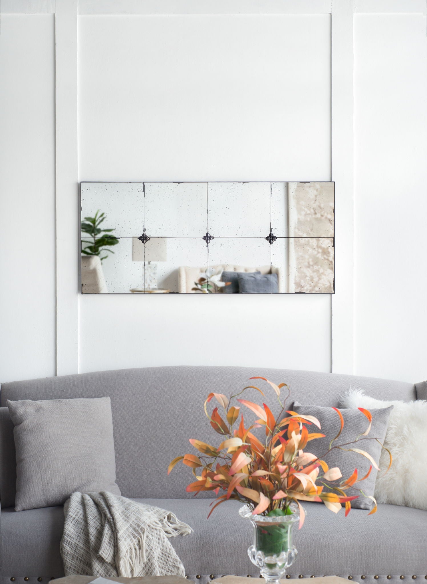 A grey sofa with neutral cushions and a beige throw sits below the Traditional Decor Style Antique Glass Panel Mirror. A glass vase with orange and green foliage decorates the coffee table in front of the sofa.