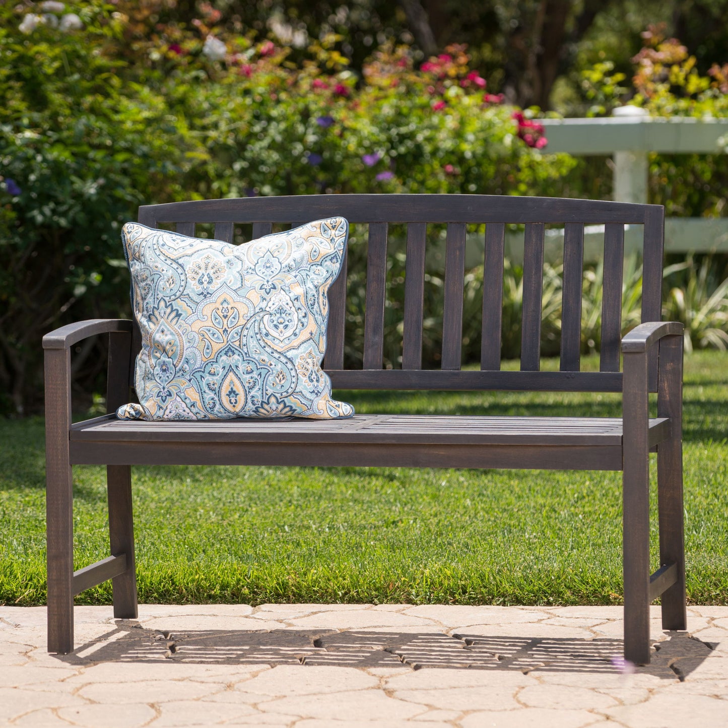 The Farmhouse Style Loja Bench Indoor / Outdoor, featuring a blue and white patterned cushion, is placed on a stone patio bordered by green grass, blooming bushes, and a white fence.