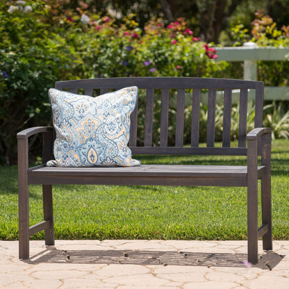 The Farmhouse Style Loja Bench Indoor / Outdoor, featuring a blue and white patterned cushion, is placed on a stone patio bordered by green grass, blooming bushes, and a white fence.