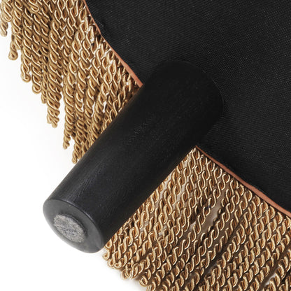 Close-up of the Yolanda - Round Upholstered Accent Ottoman, showing a black leg attached to a black fabric base with brown trim and decorative gold fringe along the edge.