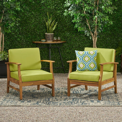 Two Perla Contemporary Club Chairs with water-resistant green cushions sit on a patterned rug on a gravel patio, backed by hedges and trees. One chair features a blue and green geometric decorative pillow.