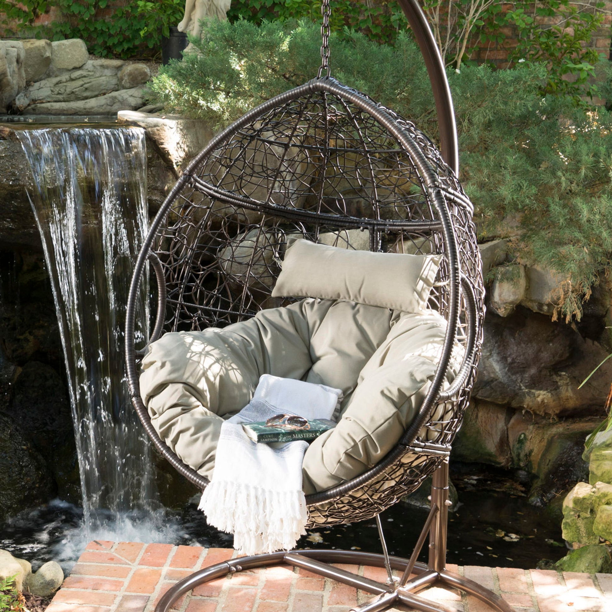 The Kylie Boho Style Hanging Basket Chair Rattan with water-resistant cushions sits on a brick patio by a pond and waterfall, surrounded by lush greenery, adorned with beige cushions and a white fringed blanket.