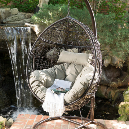 The Kylie Boho Style Hanging Basket Chair Rattan with water-resistant cushions sits on a brick patio by a pond and waterfall, surrounded by lush greenery, adorned with beige cushions and a white fringed blanket.