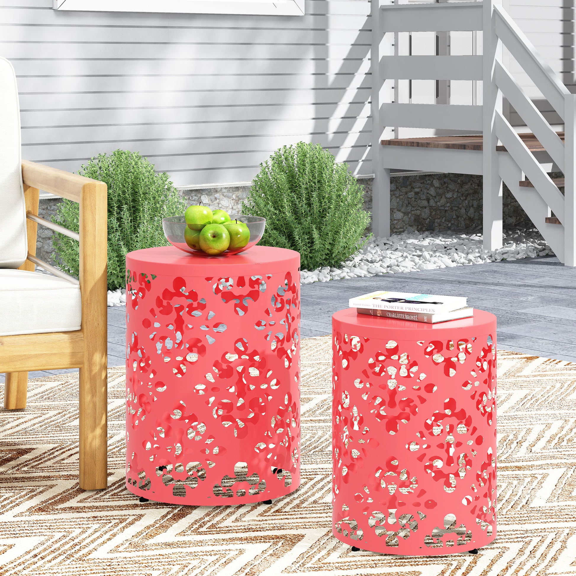 Two coral Outdoor Metal Modern Design End Tables with decorative cutouts stand on a patterned rug outdoors. One holds a bowl of green apples; the other has a book and glasses. A wooden chair and shrubs are in the background.