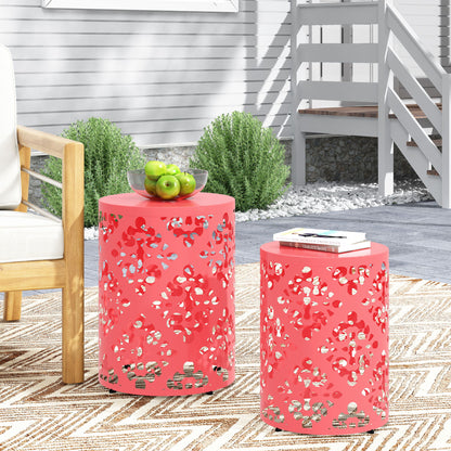 Two coral Outdoor Metal Modern Design End Tables with decorative cutouts stand on a patterned rug outdoors. One holds a bowl of green apples; the other has a book and glasses. A wooden chair and shrubs are in the background.
