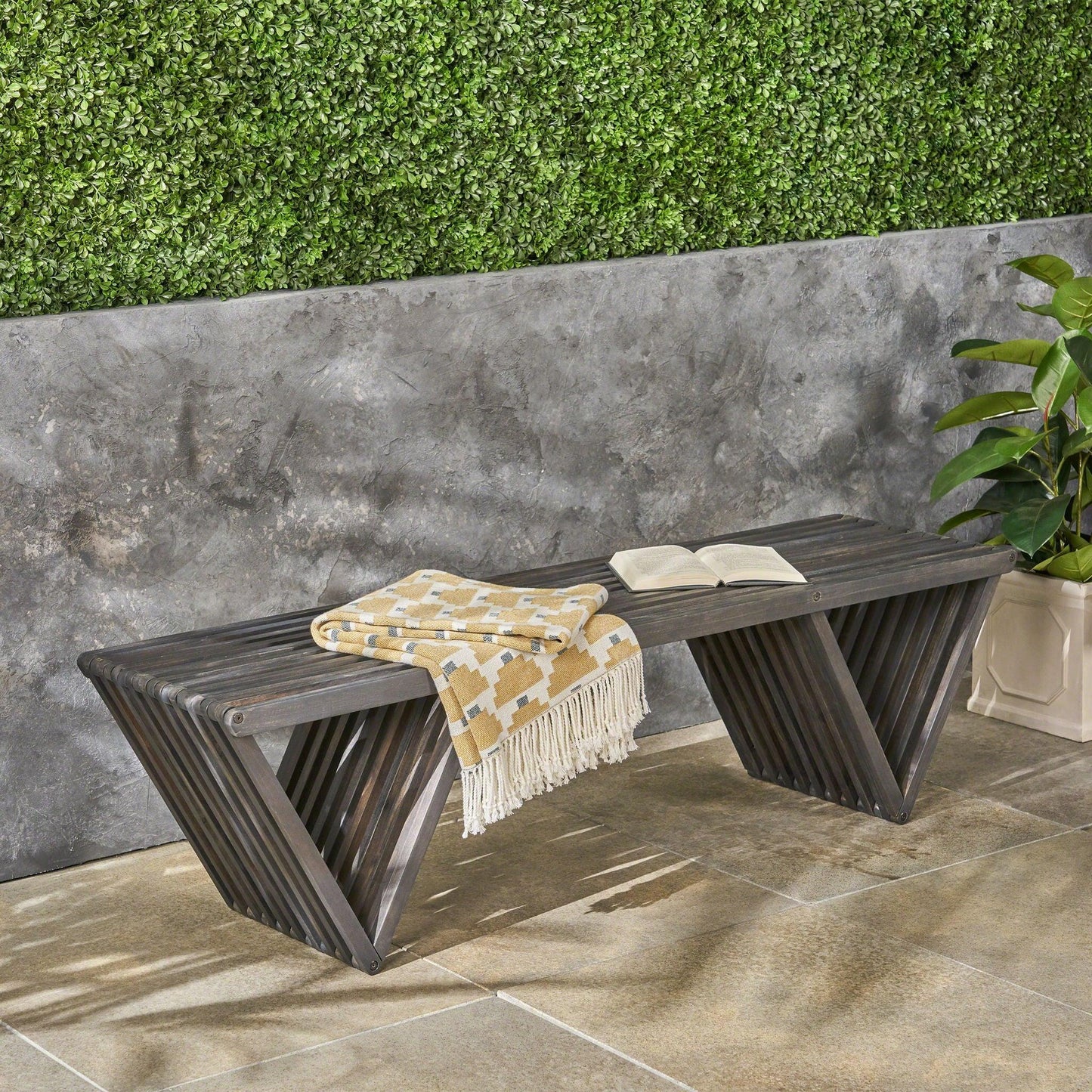 The Gloaming Outdoor Bench with a slat design and triangular base sits on a stone patio, topped with a yellow-and-white fringed blanket and an open book, set against a gray wall with a green hedge and potted plant nearby.