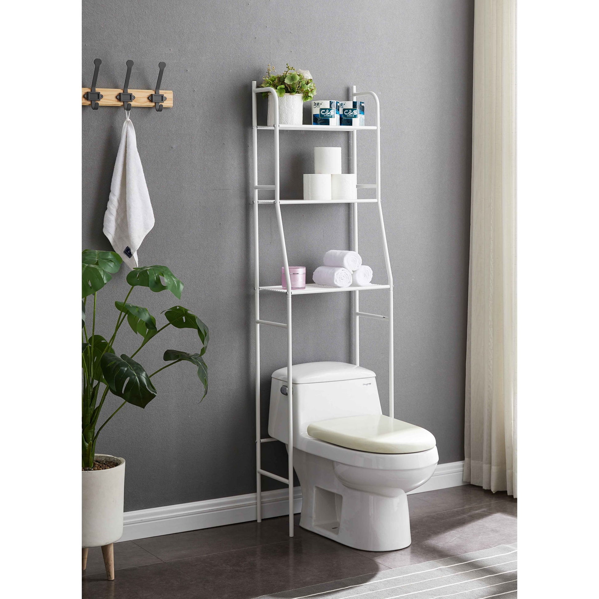 A modern bathroom features the Three Tier Over The Toilet Storage Shelf - Wood, stocked with plants, towels, and toiletries. A nearby potted plant and wall hooks with a hanging towel add style. Natural light enters through a curtain on the right.