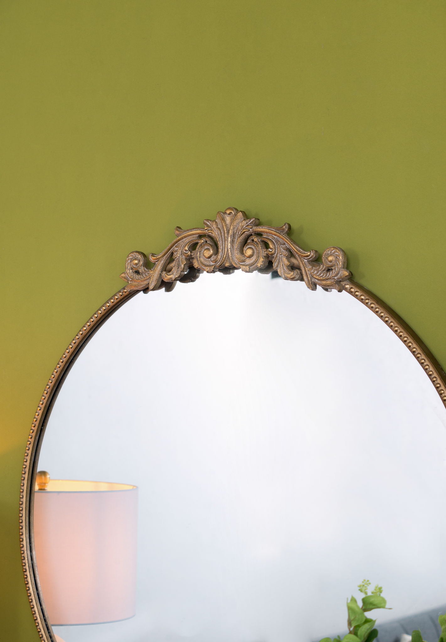 A Classic Design Mirror With Baroque Inspired Frame is mounted on a light green wall, reflecting part of a lamp and a green plant at the bottom edge.