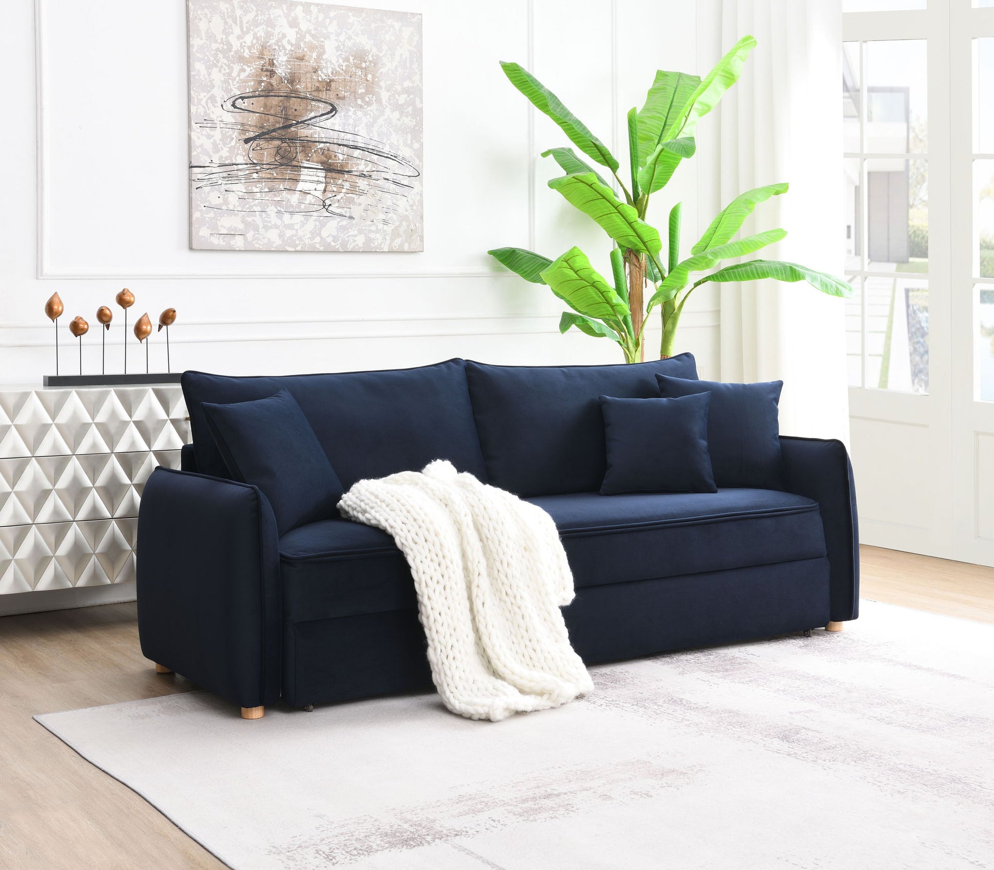 A bright, sunlit living room featuring the Irina Pull Out Sleep With Storage sofa in dark blue, topped with a white knitted blanket and matching pillows. The space is accented by a potted plant, abstract wall art, and a geometric sideboard.