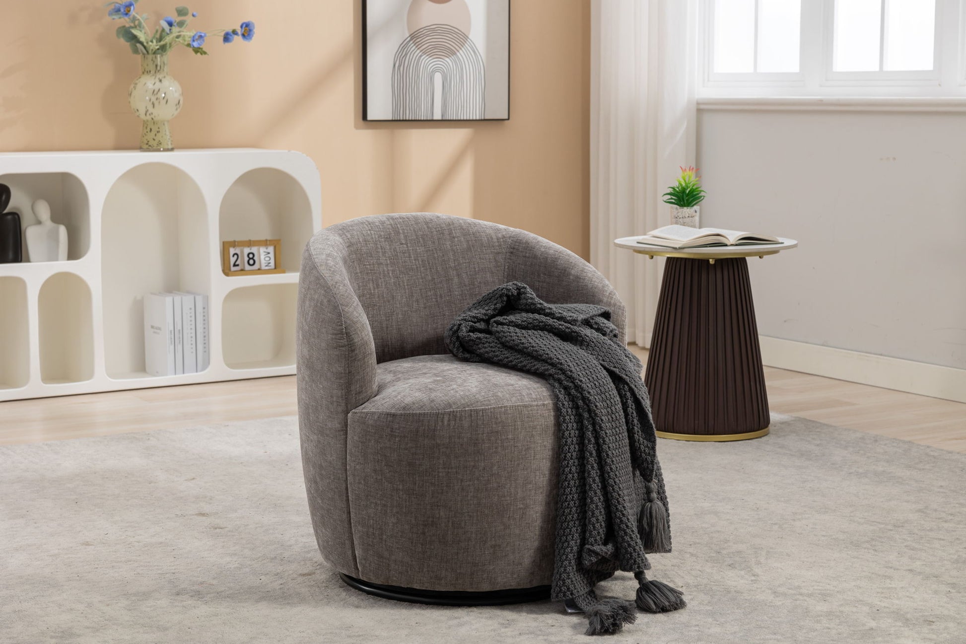 A Chenille Fabric Swivel Accent Armchair Barrel Chair with a dark knit blanket sits on a light rug. A small table nearby holds an open book and a plant, while shelves and wall art decorate the softly lit room.