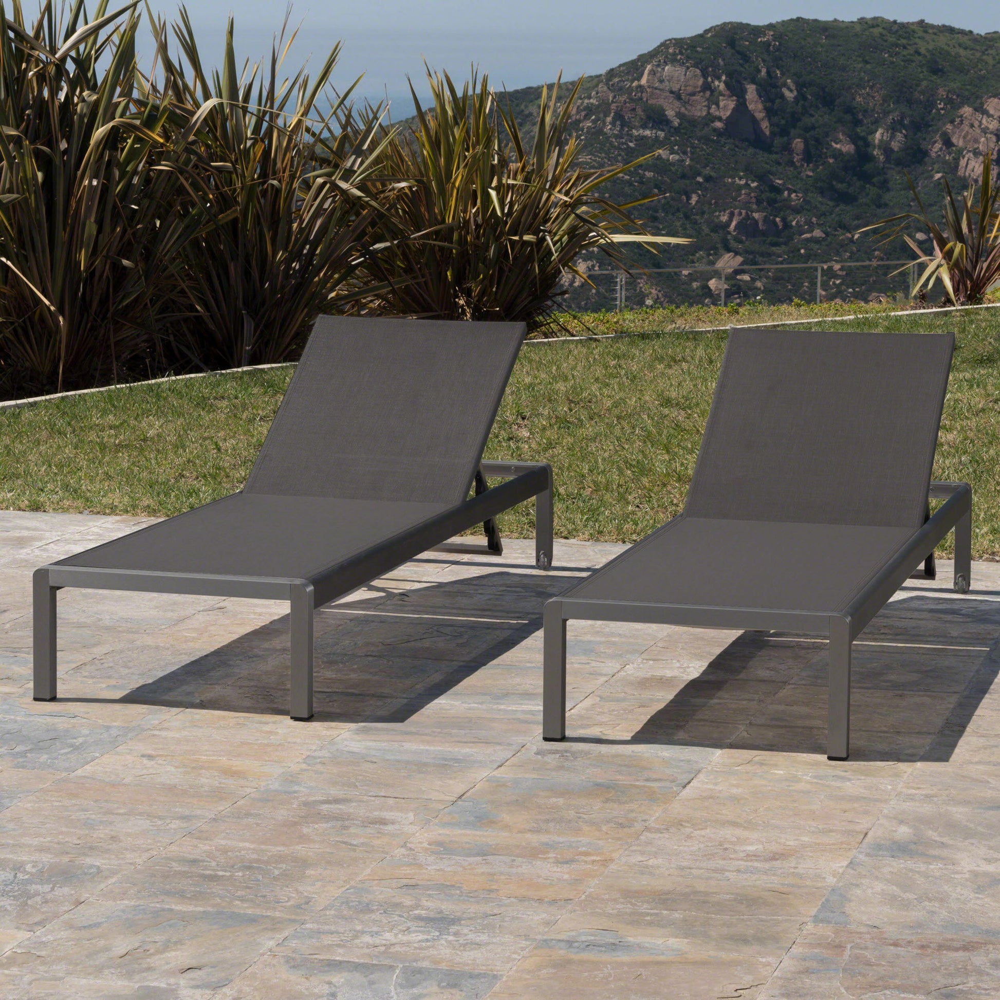 Two Outdoor Patio Chaise Lounges with 5 reclining positions are placed on a stone patio, surrounded by grass and tall plants, with mountains visible in the background under a clear sky.