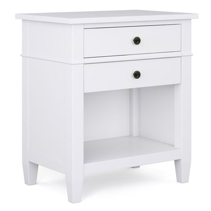The Carlton Bedside Table is a handcrafted white wooden nightstand with two drawers, round metal knobs, an open lower shelf, clean lines, and four straight legs for a simple modern look.