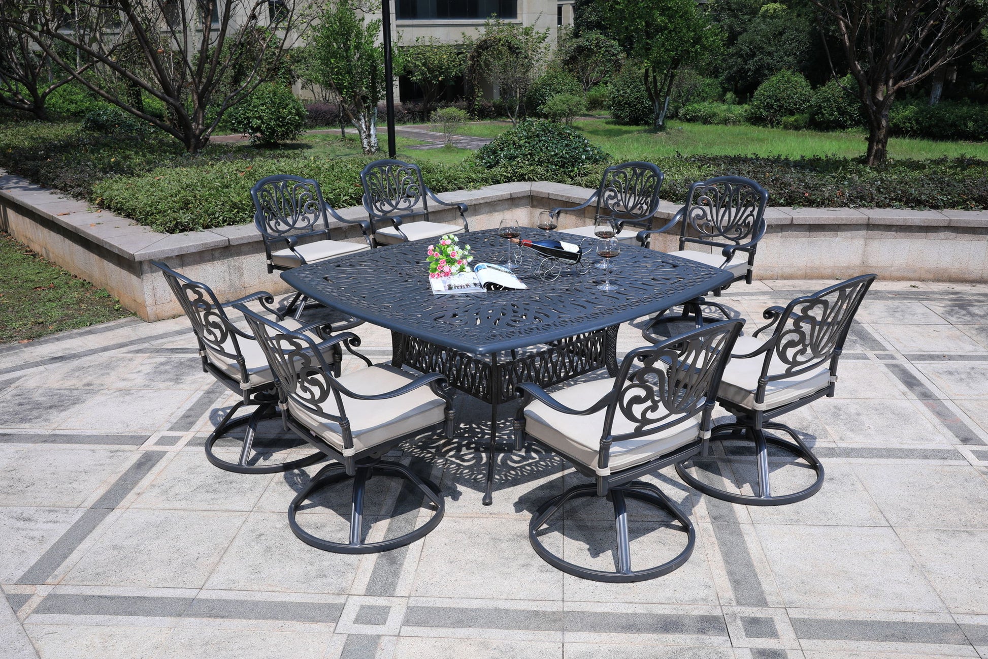 The Square 8 Person 64" Long Aluminum Dining Set With Cushions features eight matching chairs around a square table on a tiled patio, accented with flowers and small items, with lush greenery and shrubs in the background.