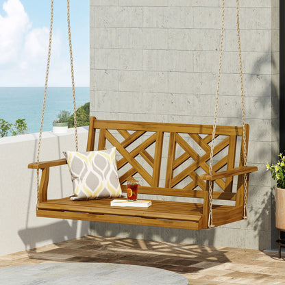 The Alejandro Acacia Wood Porch Swing with slat panel seating hangs by chains on a patio, featuring a patterned cushion, red mug, and open book on the seat. Ocean and sky are visible in the background.