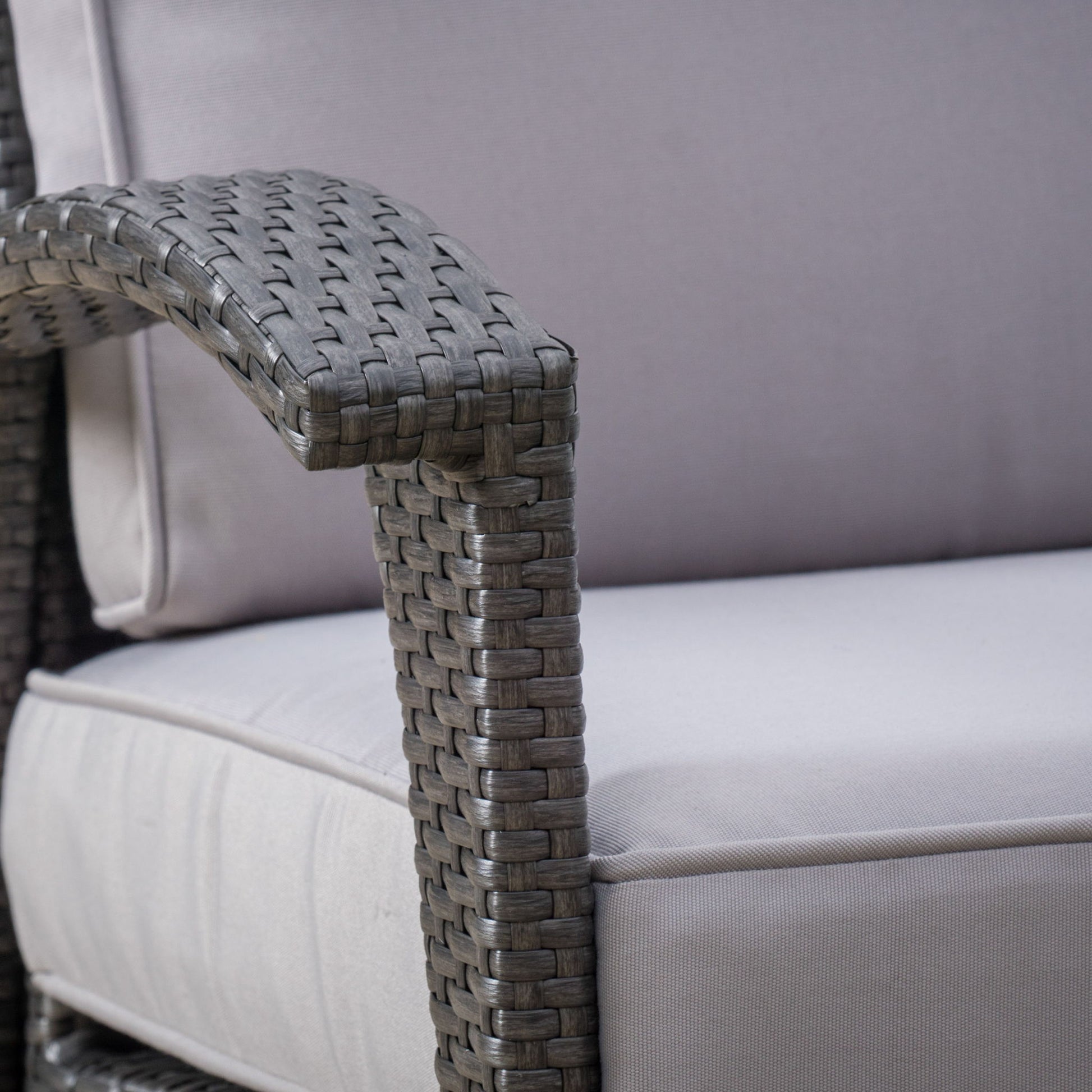 Close-up of the Honolulu - Wicker Set Outdoor Seating, highlighting the wicker armrest and light gray cushioned seat and backrest, which display the woven texture and soft fabric.