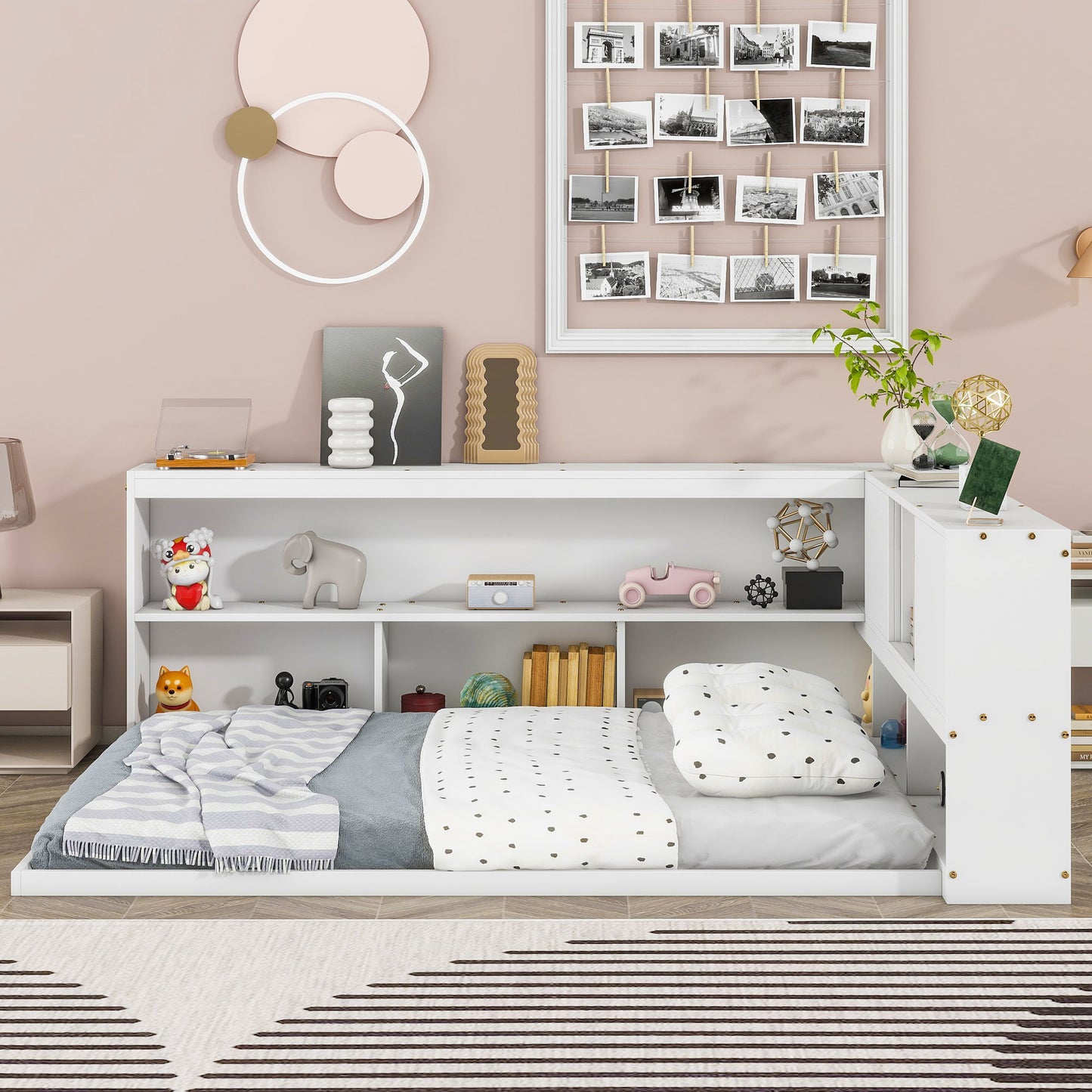 A cozy children's bedroom features a Floor Bed With L-Shaped Bookcases, Sliding Doors, Without Slats, paired with polka dot bedding, shelves for toys and books, wall art, a plant on top, light pink walls, and playful modern decor.