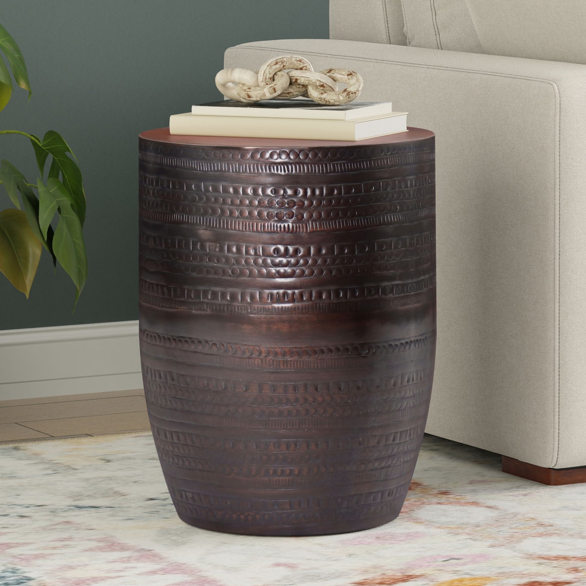 The Johnsen - Handcrafted Large Metal Accent Table, featuring dark bronze finish and textured horizontal patterns, stands beside a beige sofa. On top are stacked books and a white decor piece; nearby, a plant and patterned rug are partially visible.