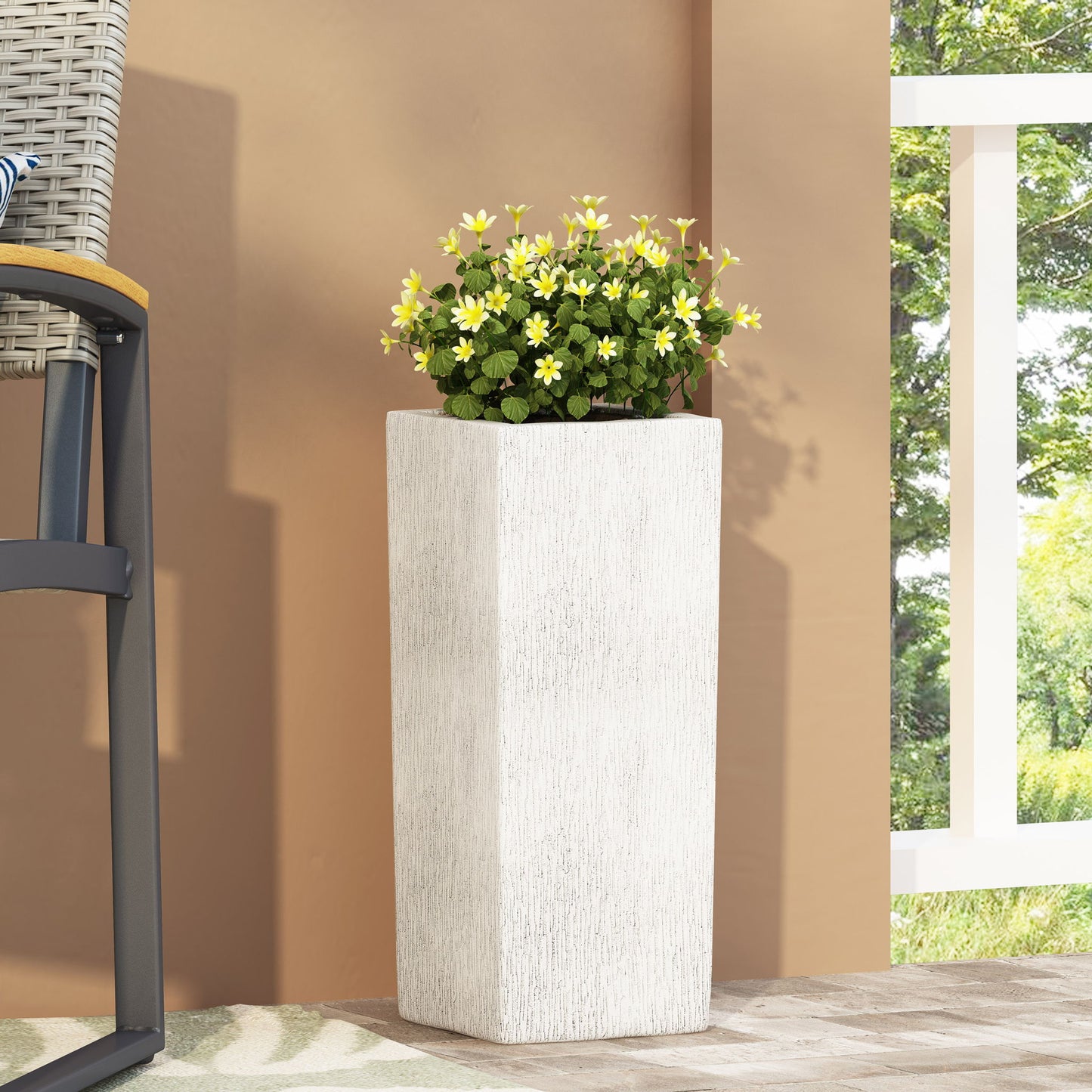 The Modern Outdoor Planter, featuring a white textured design with small yellow flowers and green leaves, is placed on a patio beside a chair, offering a glimpse of a white railing and lush greenery outside.