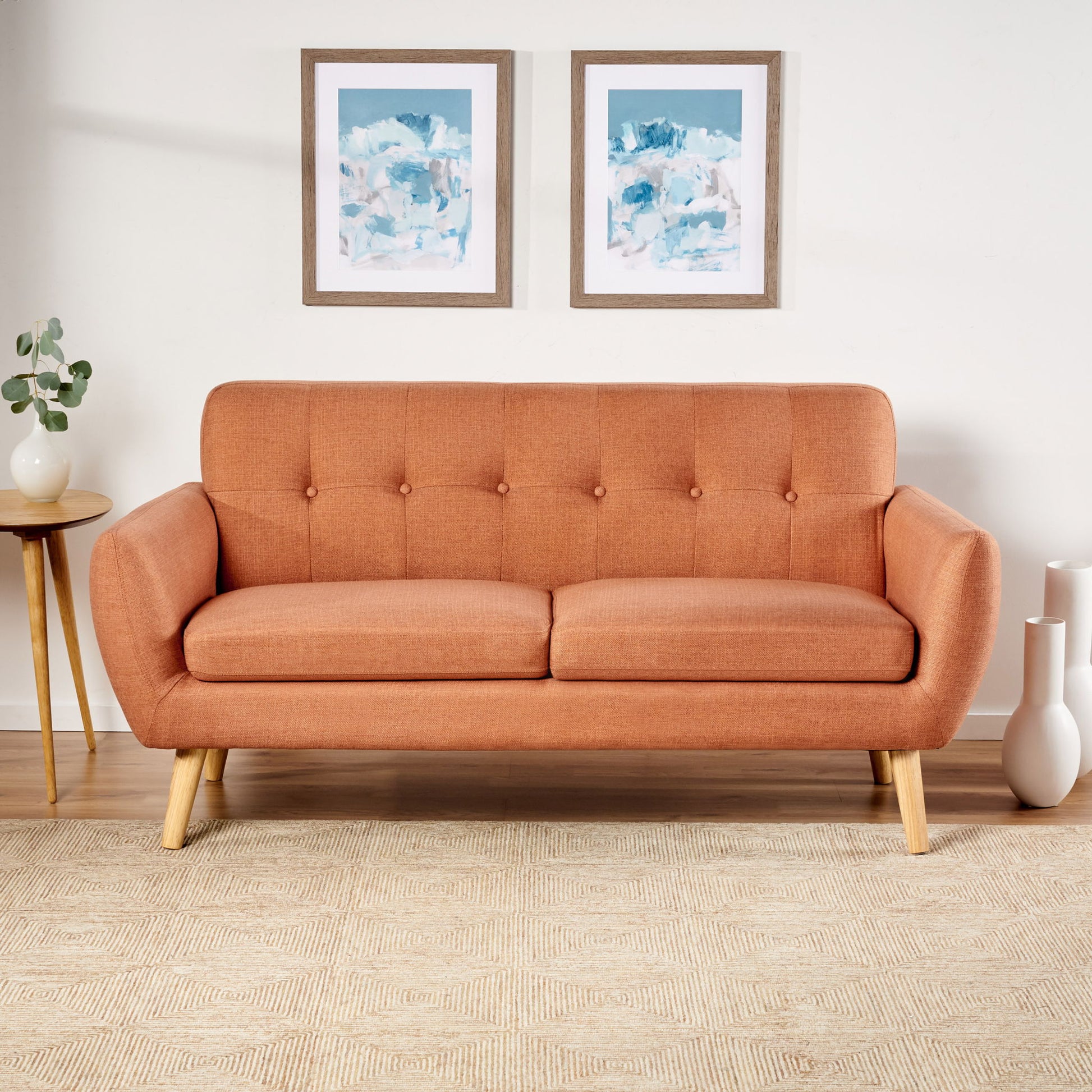 A Modern Loveseat Sofa With Button Tufted Backrest in orange with wooden legs rests on a beige rug against a white wall with two blue and white abstract artworks. Nearby, a wooden side table holds a plant and three white vases.