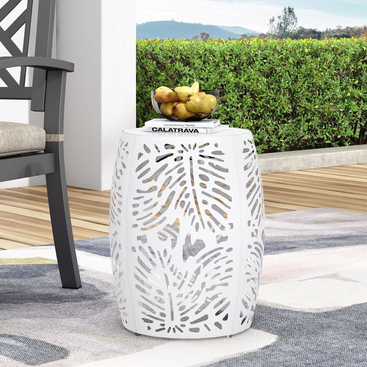 The Modern Design Metal End Table, Large, with a white finish and leaf-patterned cutouts, sits on a patio beside a chair. A stack of books and a bowl of fruit are placed on top, with green bushes and distant hills in the background.