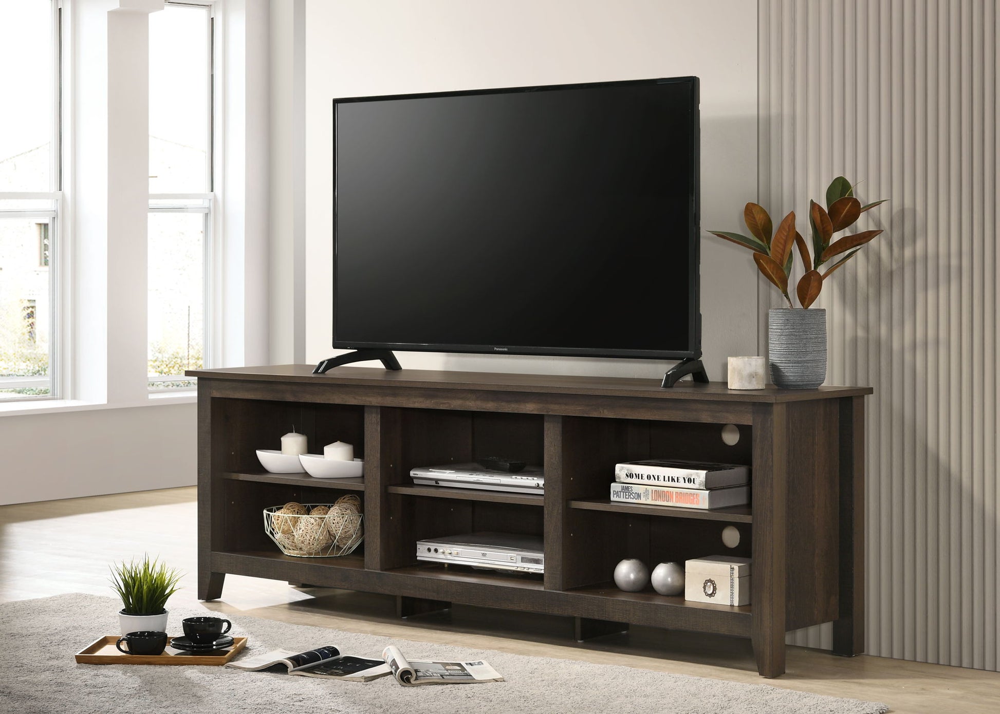 A flat-screen TV sits on the Benito - TV Stand With Open Shelves And Cable Management, which holds books, decor, baskets, and a plant in a gray vase in a bright, modern living room.