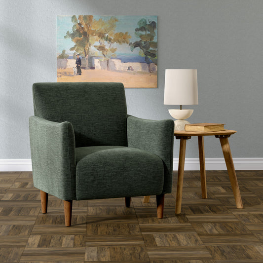 The Lexi Accent Chair in green sits beside a wooden side table with books and a white lamp on a wood-patterned floor. A landscape painting hangs on the light gray wall behind.