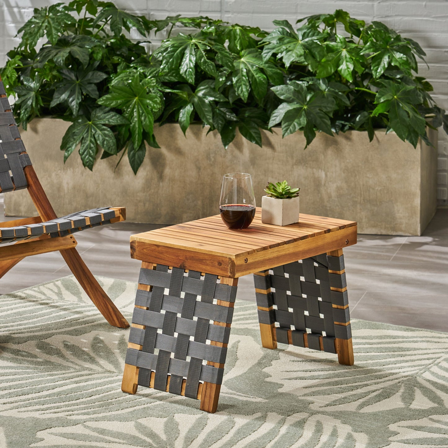 The Outdoor Wooden Foldable Side Table - Teak, featuring dark woven sides, holds a glass of red wine and a potted succulent. It stands on a patterned rug near a matching chair and large green plants in a rectangular planter.