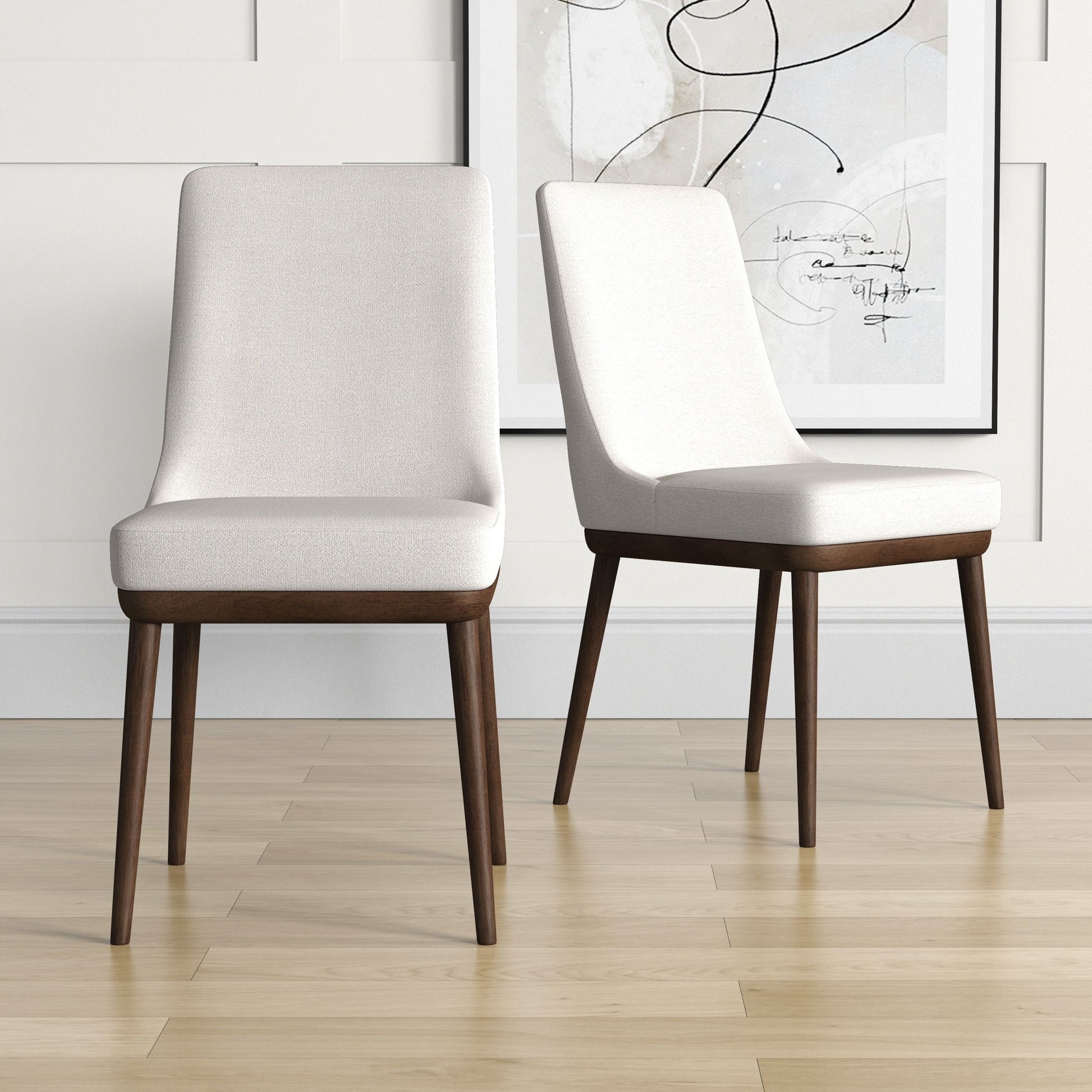 Two Kate Mid-Century Modern Dining Chairs with light upholstered seats and dark wooden legs sit on a light wood floor, with a framed abstract artwork hanging on the white paneled wall behind.