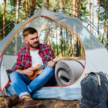 A man in a red plaid shirt plays a ukulele outside his tent in the forest. Nearby, a Memory Foam Camping Mattress with water-resistant cover and a backpack rest on the ground among tall trees.