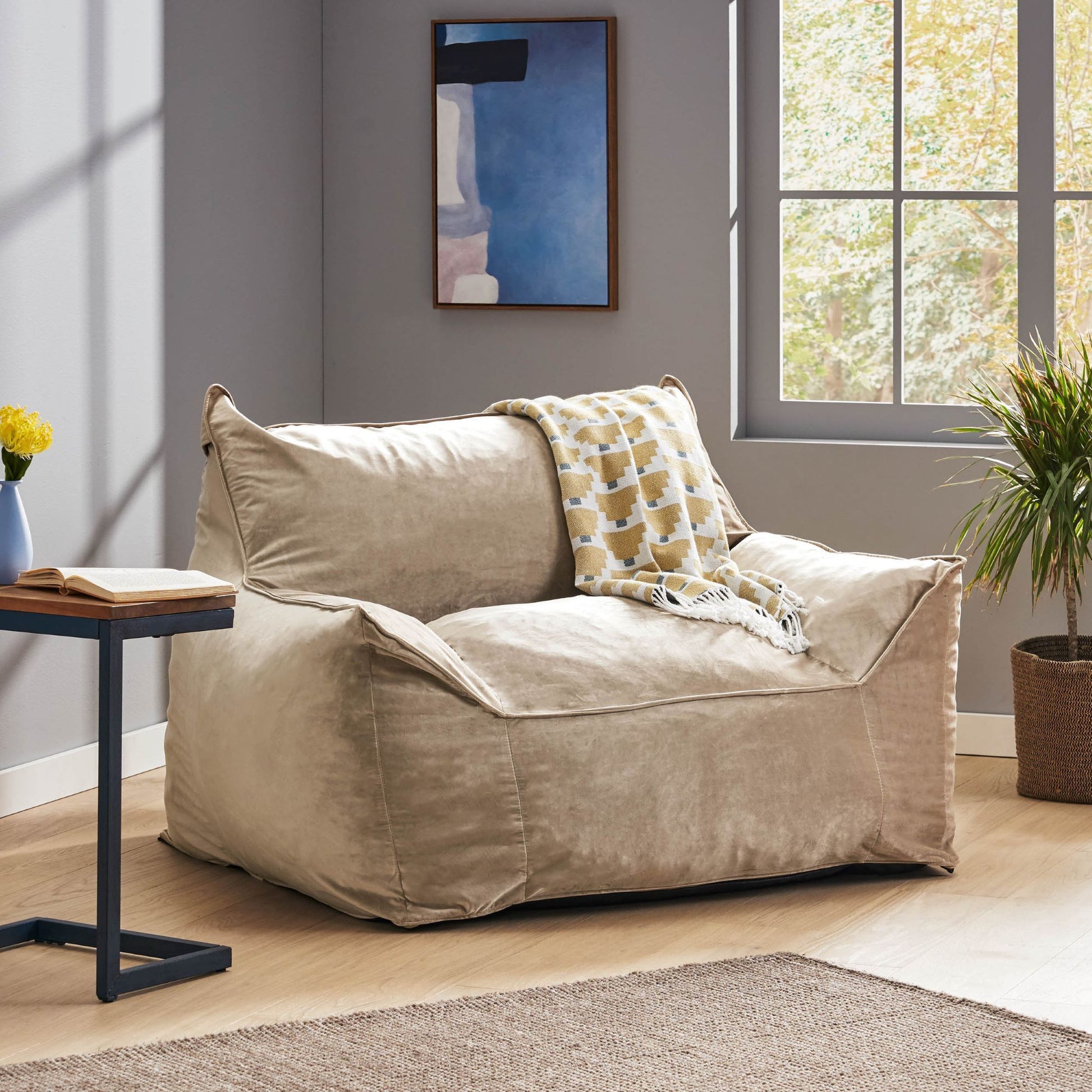 The Allea - Bean Bag Chair With Armrests in beige sits in a sunlit room by the window, accompanied by a patterned throw blanket, a small table, a potted plant, and modern wall art.