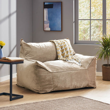 The Allea - Bean Bag Chair With Armrests in beige sits in a sunlit room by the window, accompanied by a patterned throw blanket, a small table, a potted plant, and modern wall art.