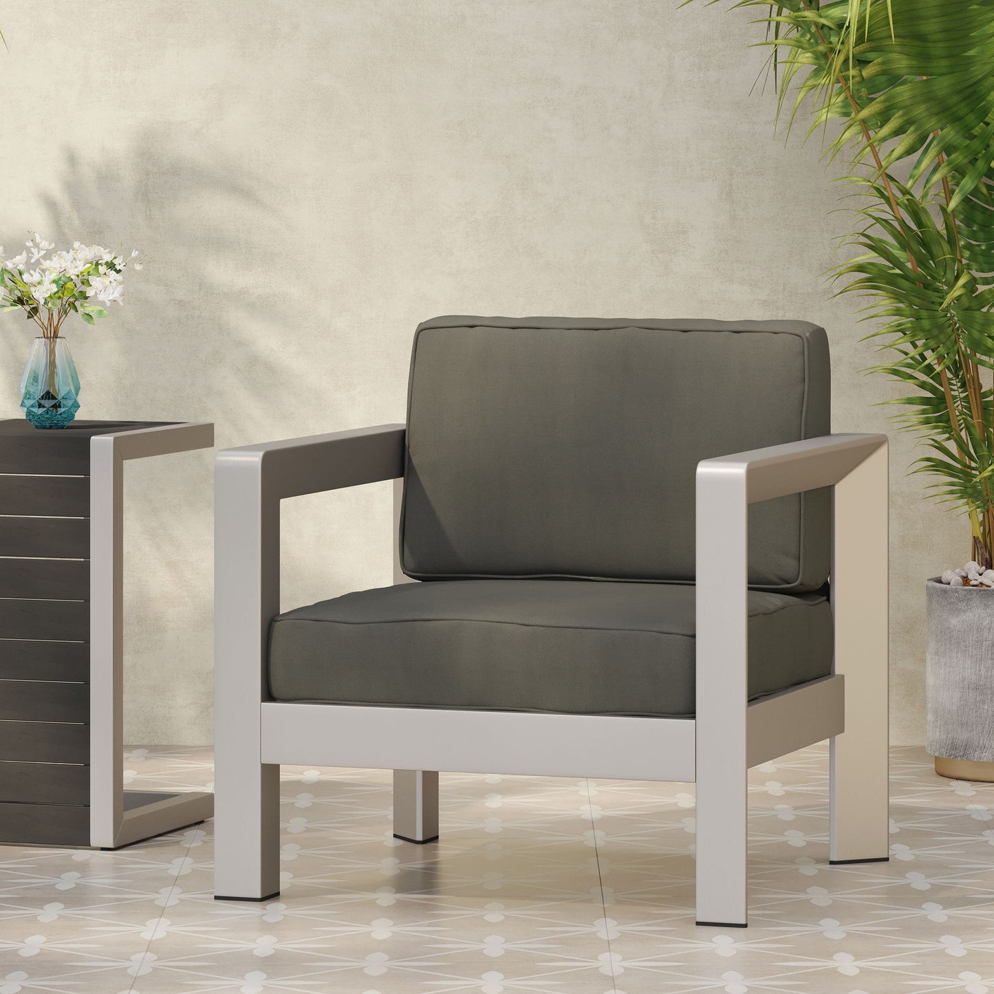 The Aviara - Luxurious Outdoor Club Chair with gray cushions and a silver metal frame sits on tiled flooring beside a black side table holding a small vase of flowers, with green plants against a light textured wall in the background.