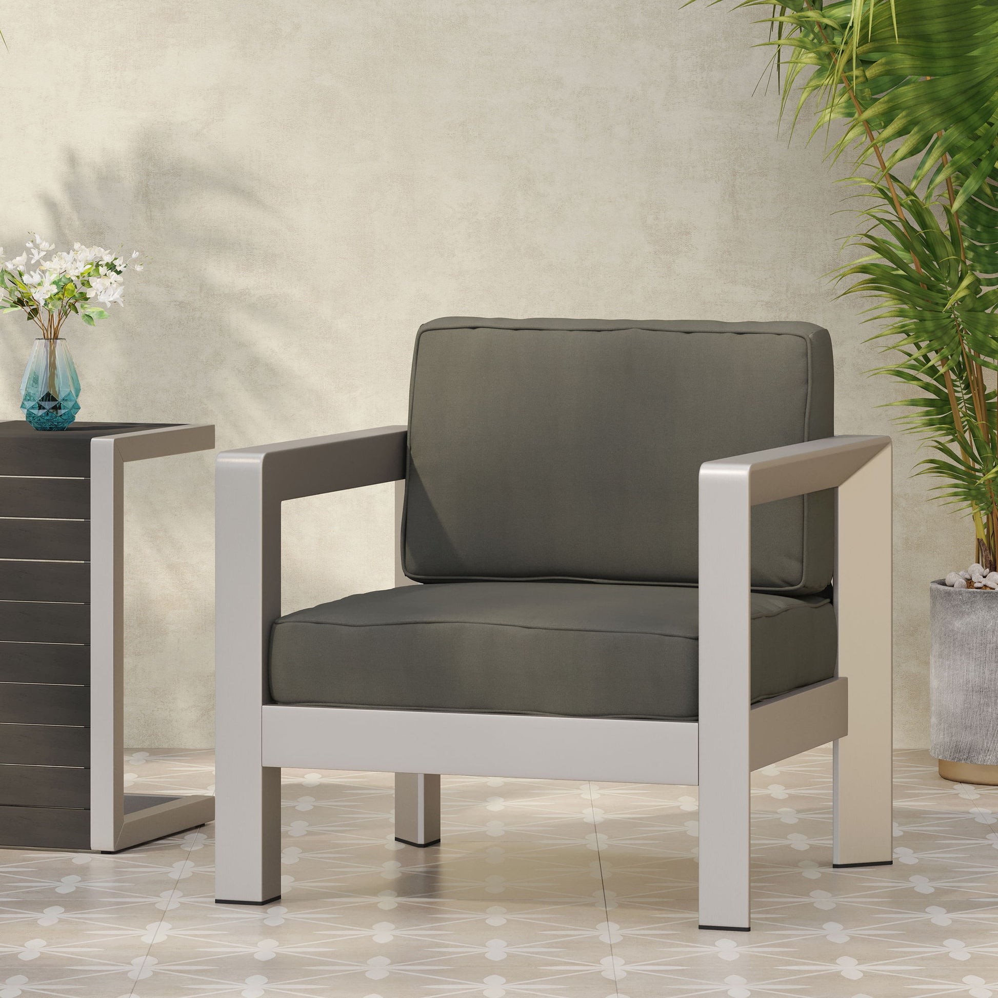 The Aviara - Luxurious Outdoor Club Chair with gray cushions and a silver metal frame sits on tiled flooring beside a black side table holding a small vase of flowers, with green plants against a light textured wall in the background.