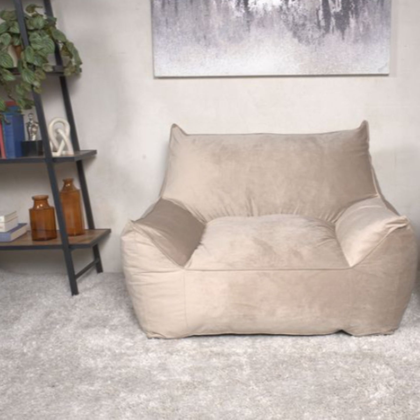 The Allea - Bean Bag Chair With Armrests in beige sits cushioned on a light gray rug in a modern living room. To the left is a bookshelf with plants and decor, while a neutral abstract painting hangs behind the chair.