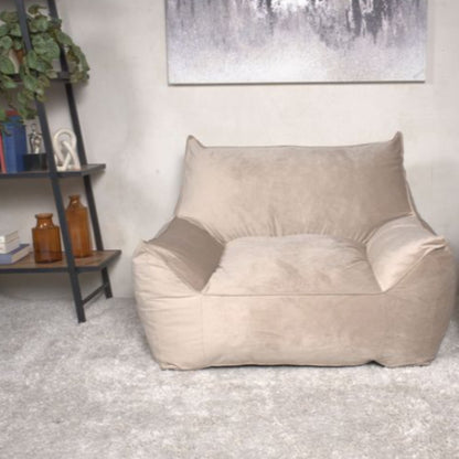 The Allea - Bean Bag Chair With Armrests in beige sits cushioned on a light gray rug in a modern living room. To the left is a bookshelf with plants and decor, while a neutral abstract painting hangs behind the chair.