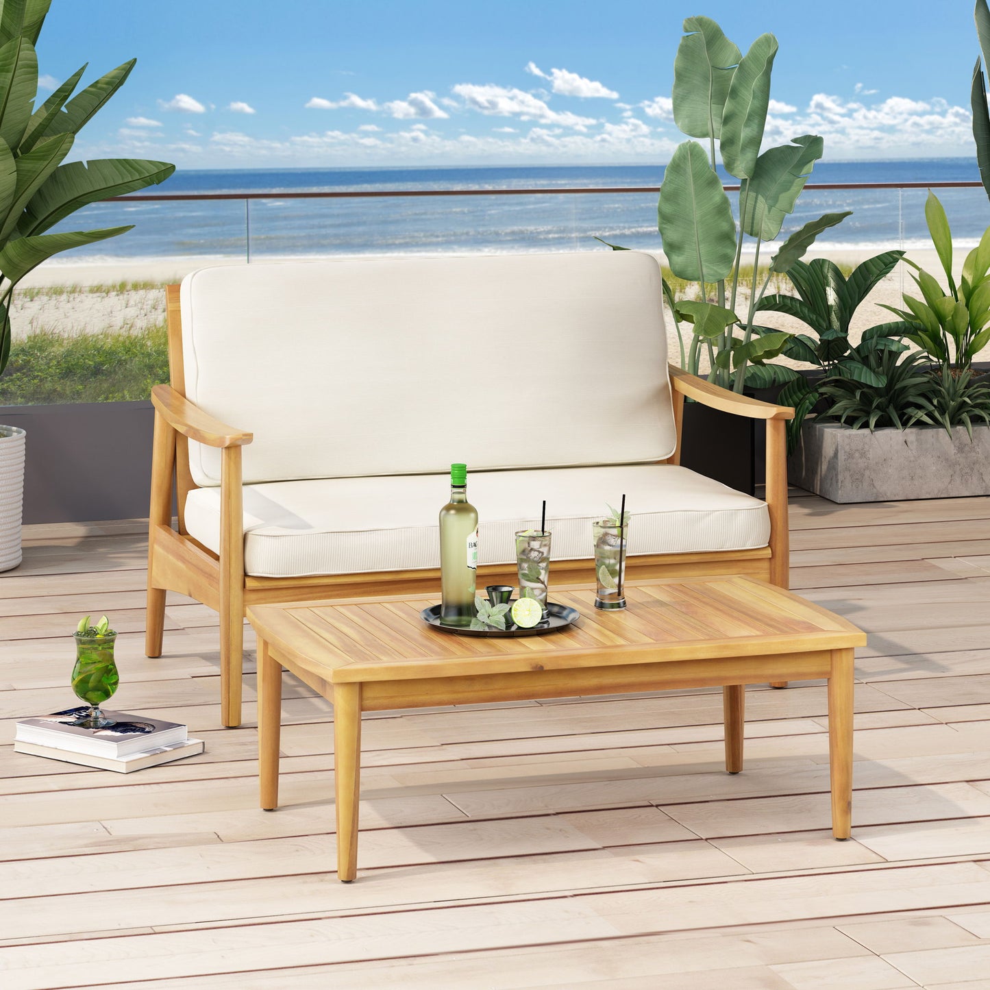 The Outdoor Acacia Wood Loveseat Set With Coffee Table features a wooden loveseat with white cushions and a matching table holding drinks, set on a deck with green plants and an ocean view in the background.