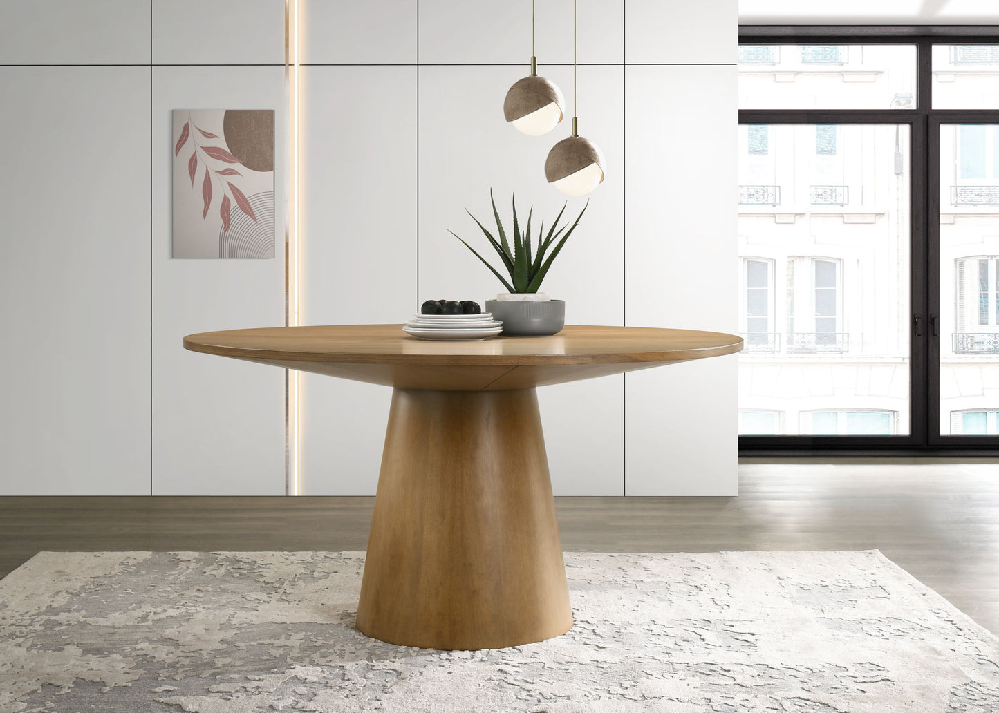 The Jasper - Wide Contemporary Round Dining Table stands on a patterned rug, topped with a potted plant and figs. Modern pendant lighting hangs above, while large windows and minimal white decor complete the contemporary setting.