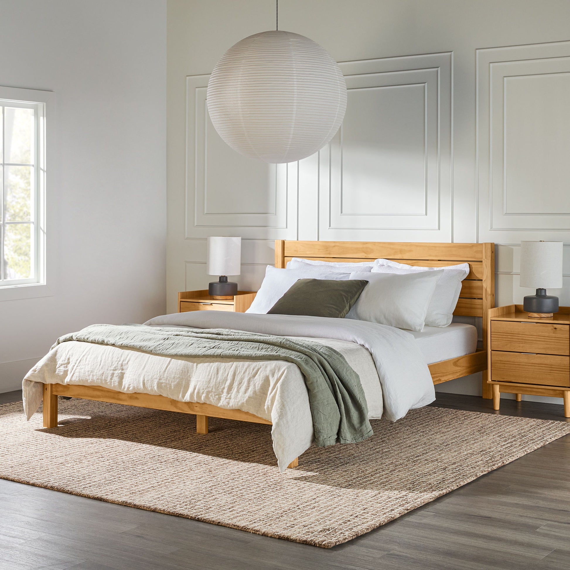 A modern bedroom features the Modern Minimalist Boho Bedframe, white and green bedding, two wooden nightstands with lamps, a large round paper lantern overhead, natural window light, and a textured rug on the floor.