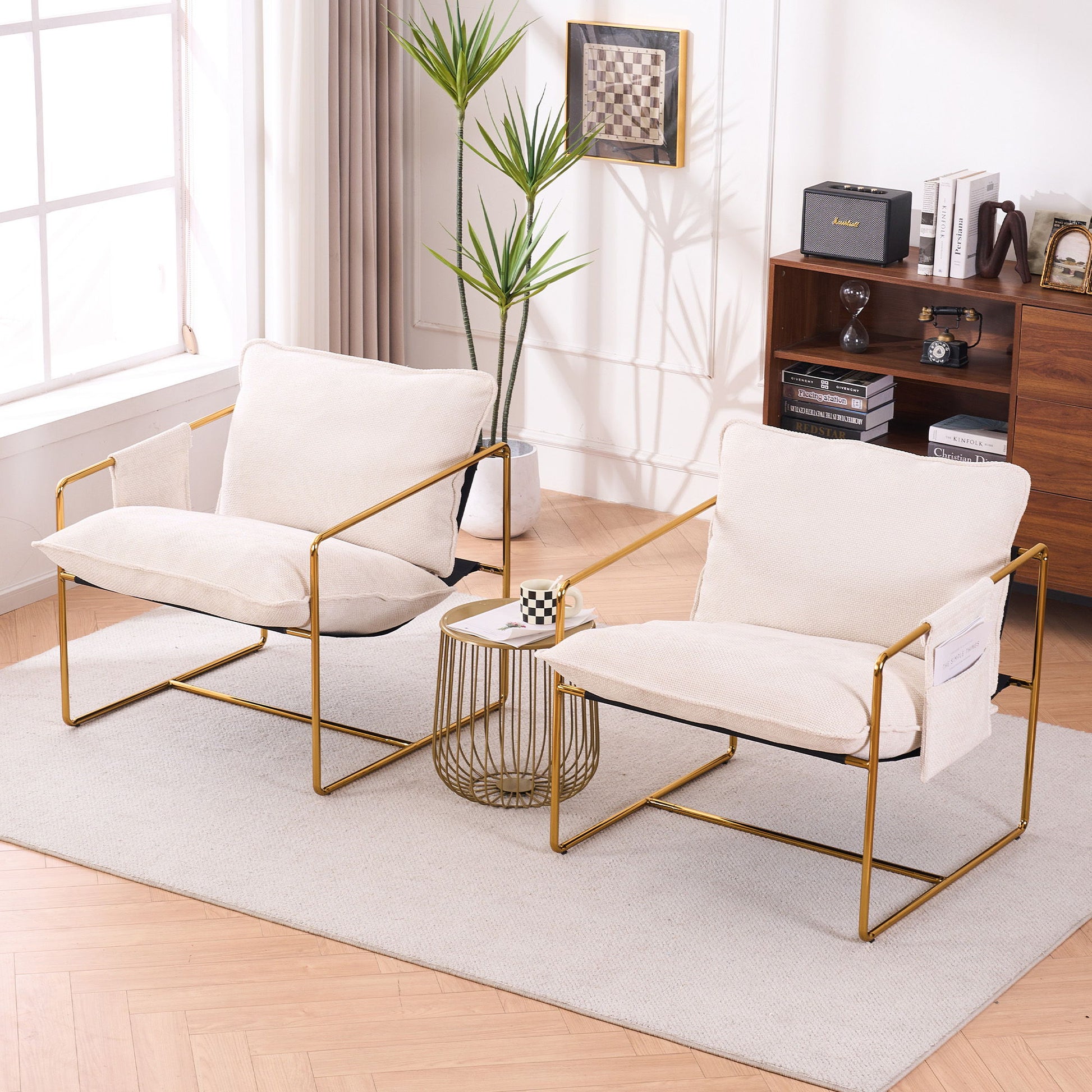 Two Upholstered Hanging Armchairs with gold-plated metal frames, crushed foam cushions, and skin-friendly fabric sit on a rug by a small table with a chessboard, backed by a mid-century bookshelf and a tall plant near the window.