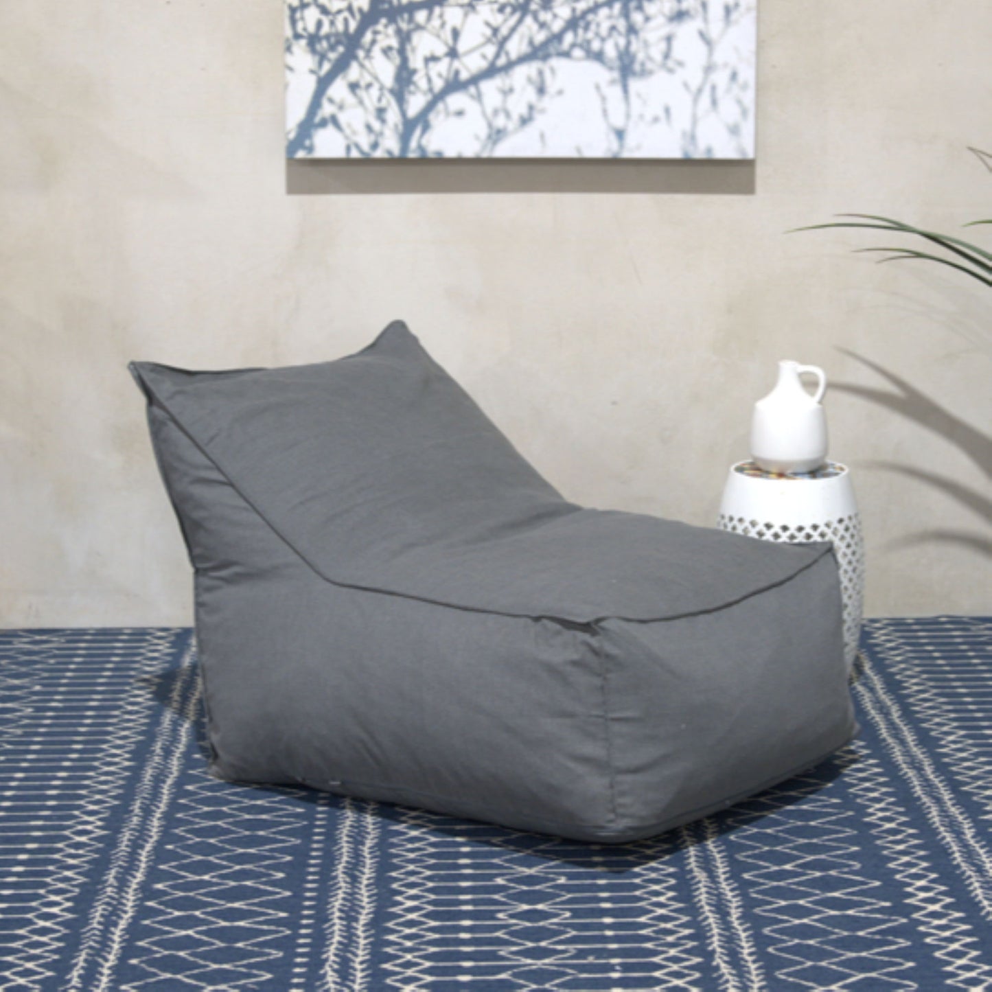 The Ettie Water Resistant Fabric Bean Bag Chair in gray sits on a patterned blue rug beside a white side table with a white pitcher, while wall art and green leaves appear in the background.